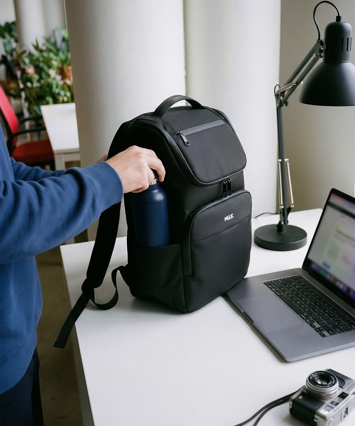 Person placing a water bottle into a black backpack on a desk with a laptop and camera.