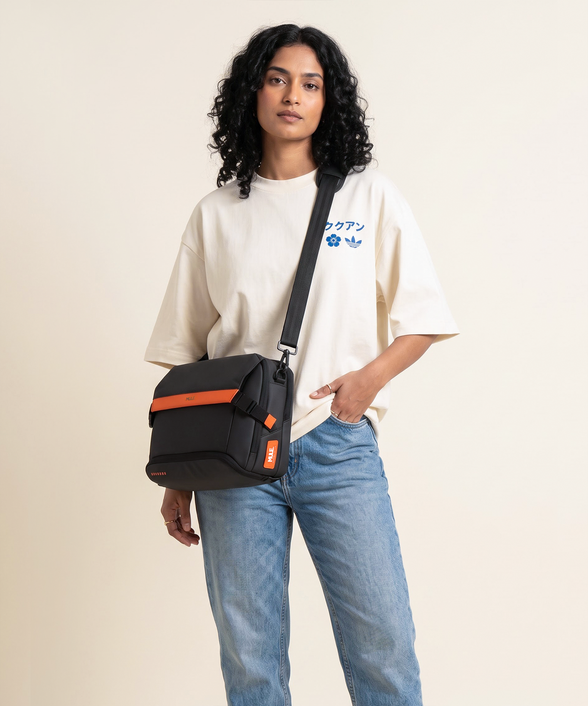 A model wearing a Mule hatch messenger bag with orange accents and a black strap, styled over a cream graphic t-shirt and blue jeans against a solid beige background.