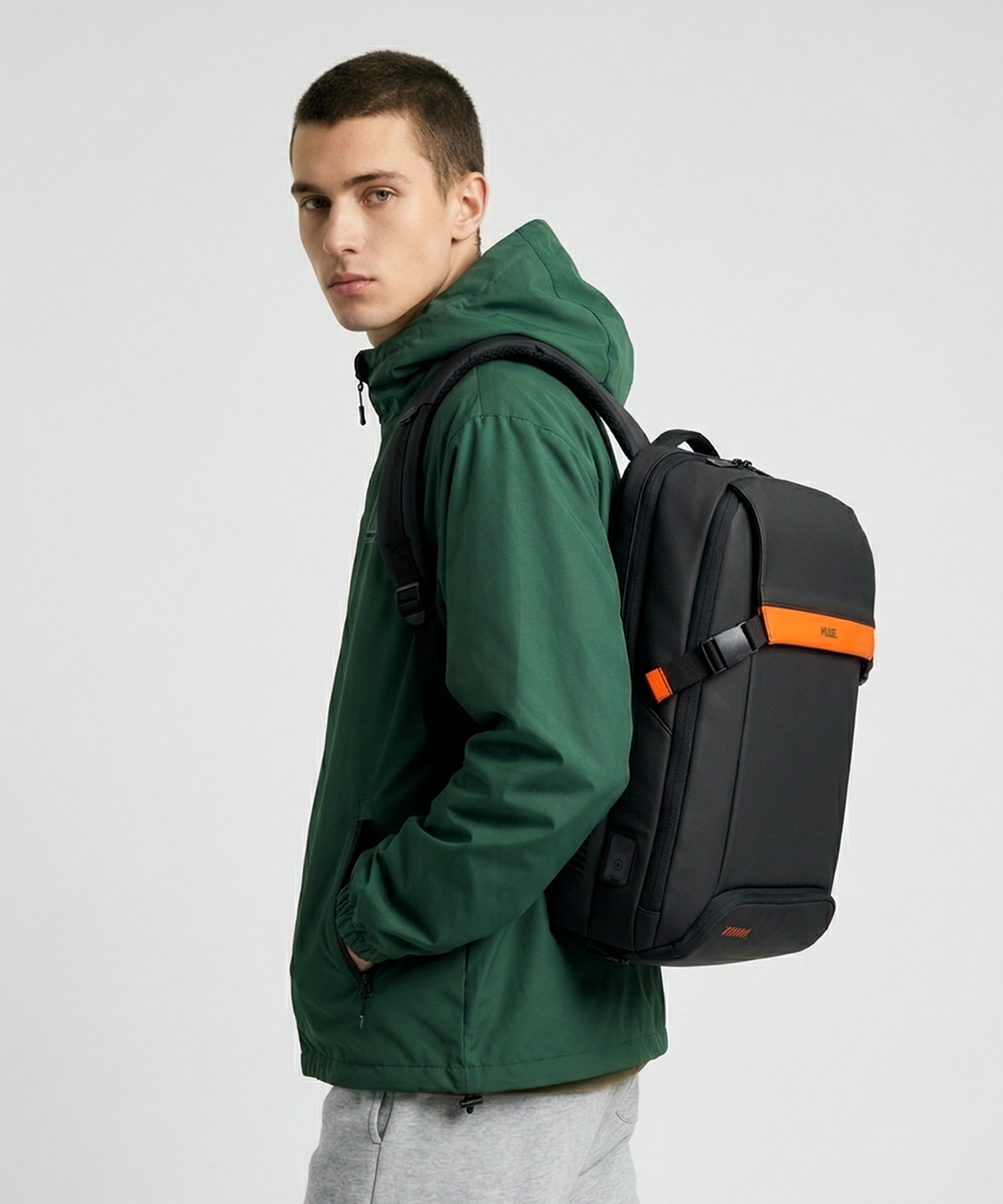 A man wearing a dark green hooded jacket and gray sweatpants, shown from the side/back, wearing a MULE hatch backpack with a bright orange horizontal stripe and orange logo on the front, against a light gray studio background.