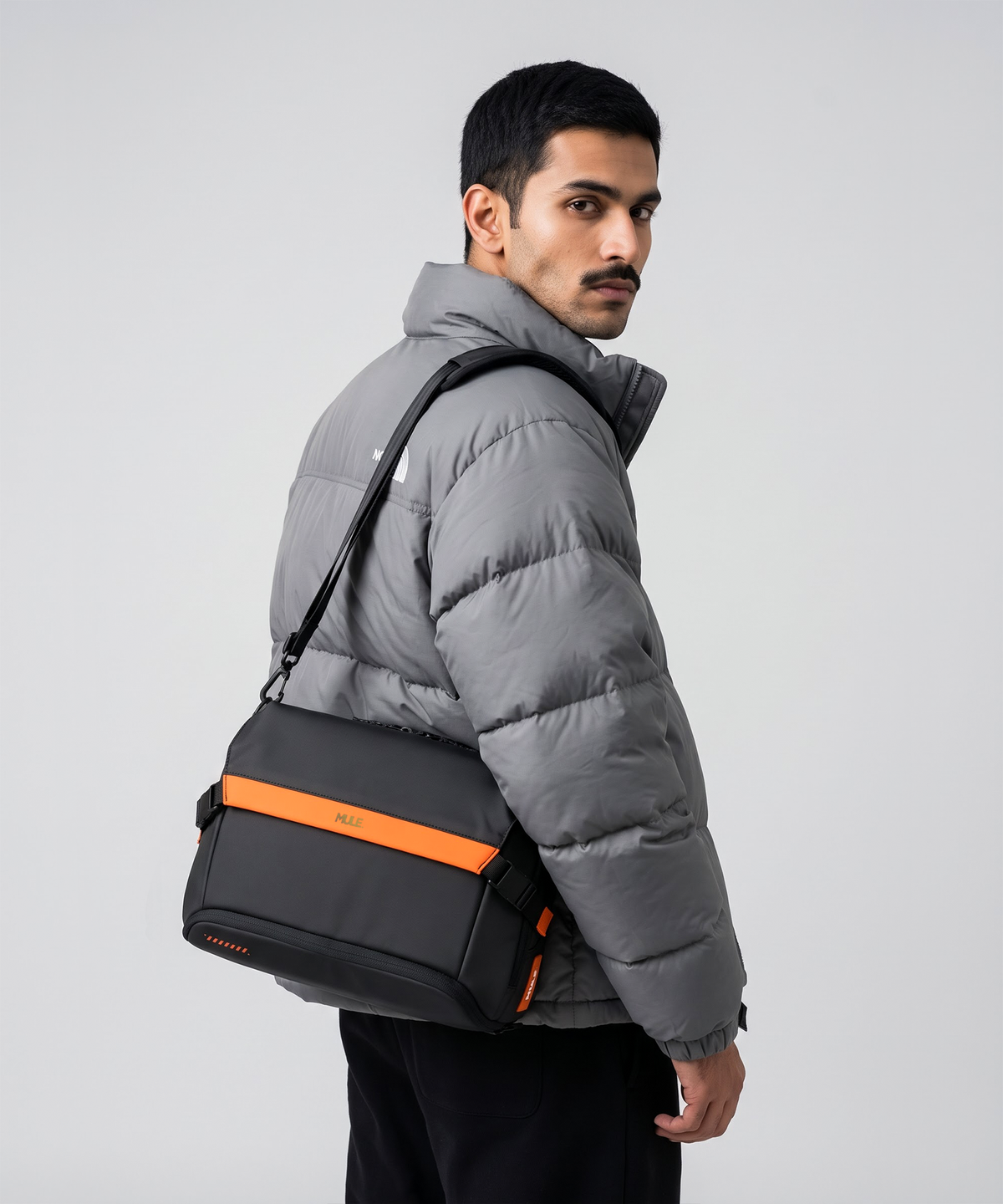 A man carries a black messenger bag with a bright orange accent stripe and the brand name 'Mule' on the front, set against a plain light gray studio background.