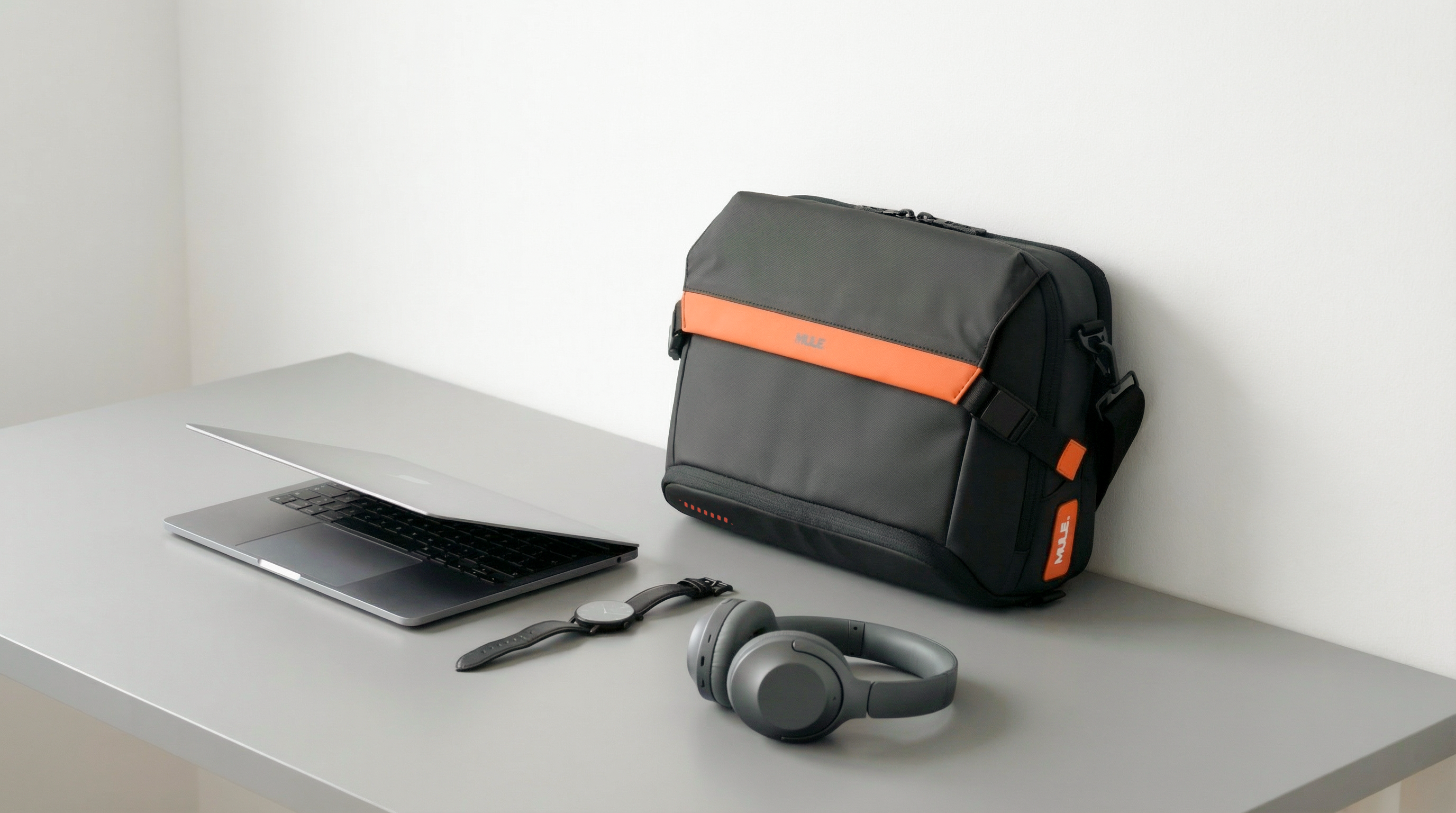 Black messenger bag with orange accents on a white surface next to a laptop, keyboard, and headphones.