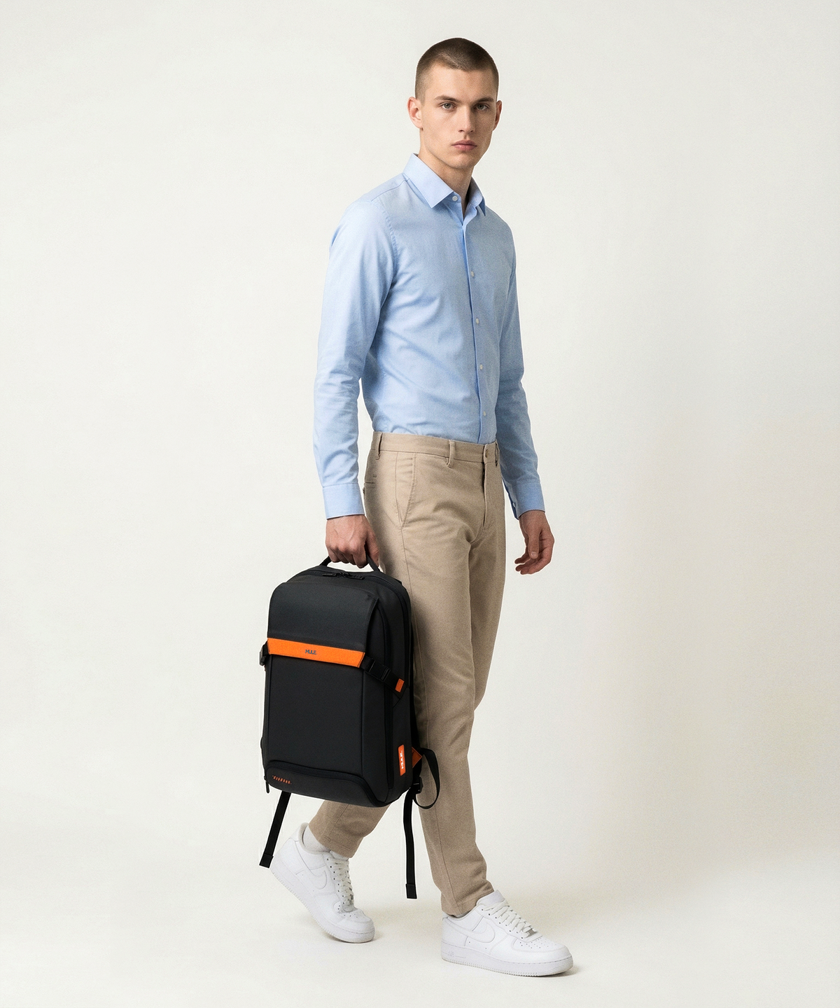 A man wearing a light blue dress shirt, beige trousers, and white sneakers holds a black Hatch backpack with an orange accent stripe against a plain light beige studio background.