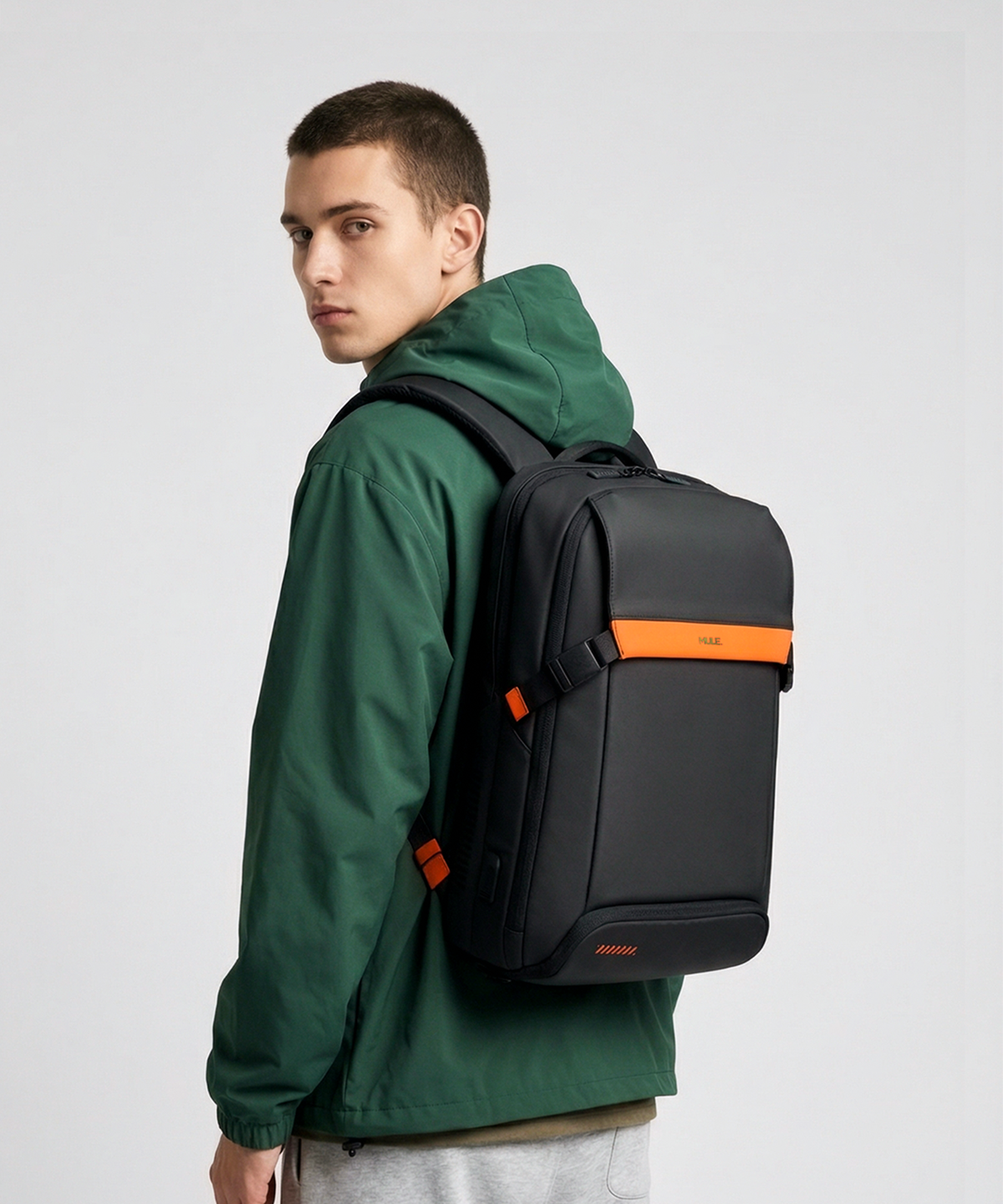 A man wearing a dark green hooded jacket and gray sweatpants, shown from the side/back, wearing a MULE hatch backpack with a bright orange horizontal stripe and orange logo on the front, against a light gray studio background.