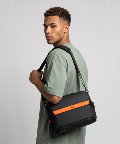 A man wearing a sage green t-shirt carries a black messenger bag with a bright orange accent stripe and the brand name 'Mule' on the front, set against a plain light gray studio background.