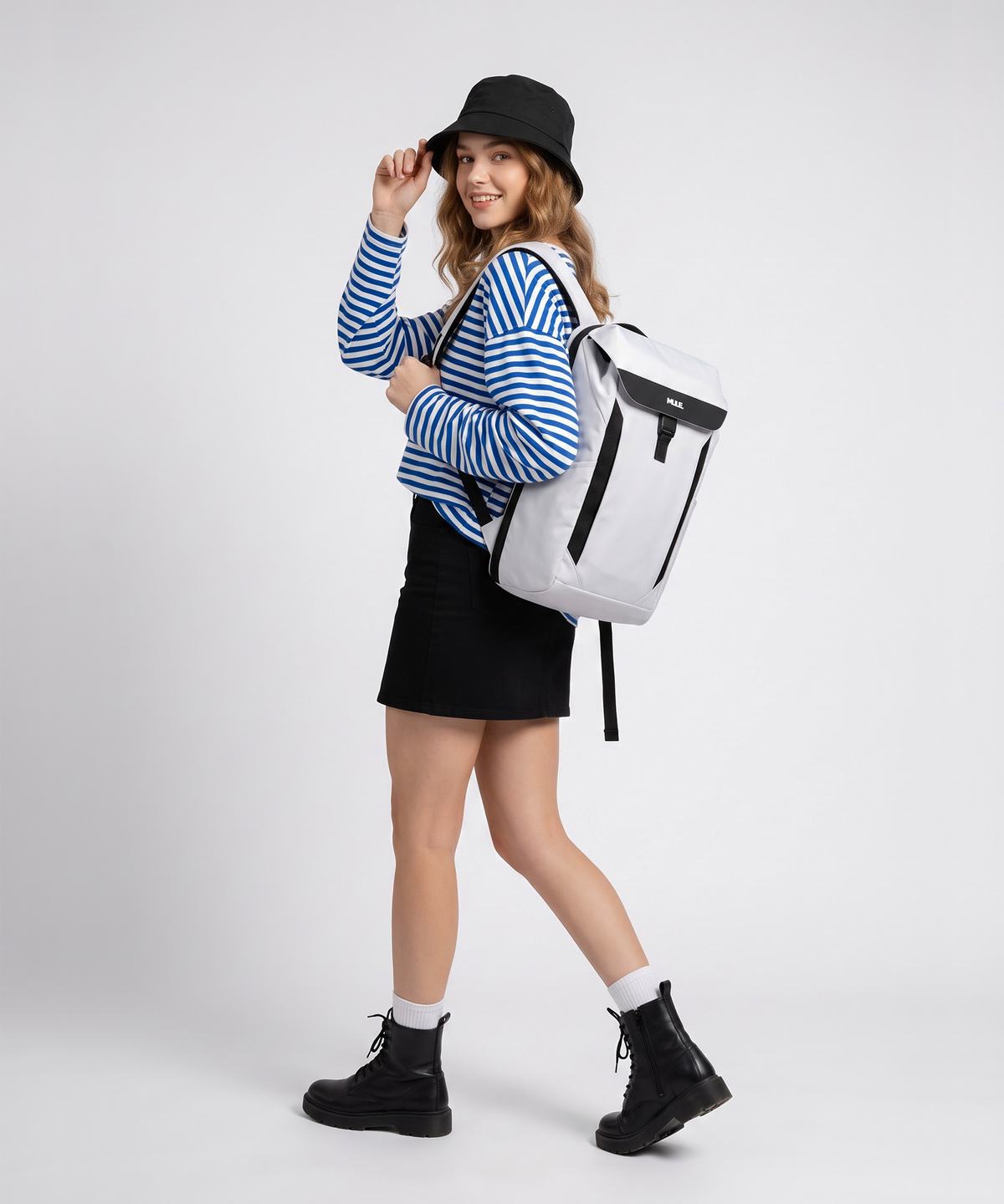 A woman modelling a white Mule Kepler backpack with black accents, styled with a blue and white striped long-sleeve shirt, a black mini skirt, a black bucket hat, and black combat boots against a light grey studio background.