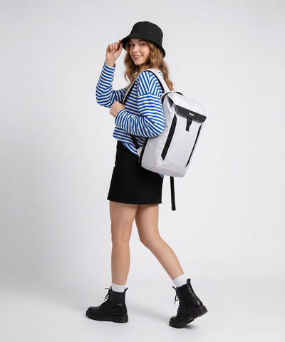 A woman modelling a white Mule Kepler backpack with black accents, styled with a blue and white striped long-sleeve shirt, a black mini skirt, a black bucket hat, and black combat boots against a light grey studio background.