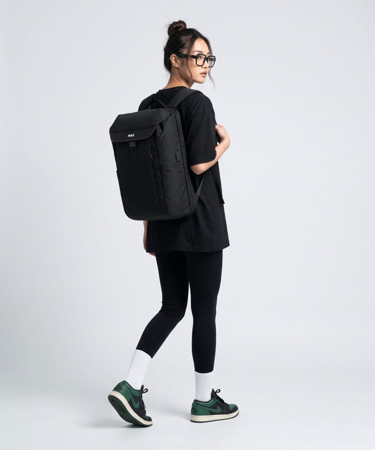 A woman wearing a black t-shirt, black leggings, and green sneakers carries a black Mule backpack with a top flap and front zipper pocket, photographed against a light gray studio background.