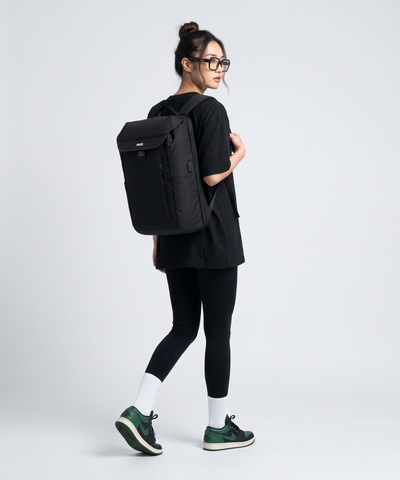 A woman wearing a black t-shirt, black leggings, and green sneakers carries a black Mule backpack with a top flap and front zipper pocket, photographed against a light gray studio background.